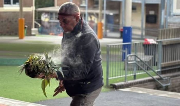 An Aboriginal man holding a smoking leaves doing a smoking ceremony in a school playground