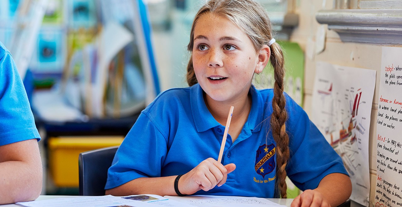 Young female student looks up while sitting at desk and holding a pencil over an open notebook 