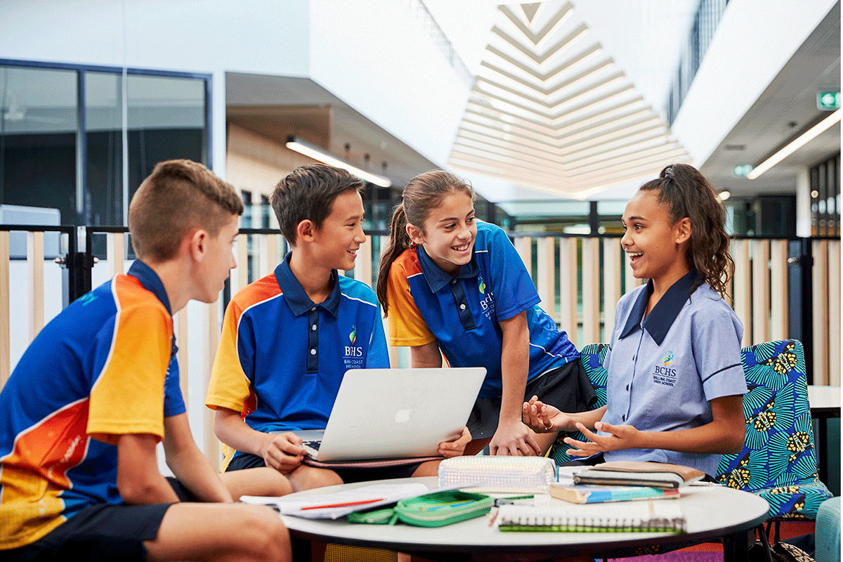 An animated group of high school students talking at a round desk with computer and workbooks