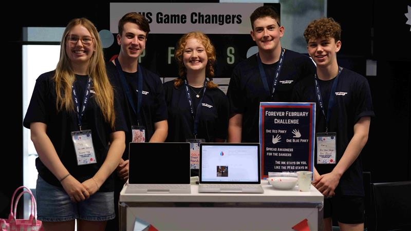 All 5 students smiling, standing next to each other with 2 laptops on the table in front of them.