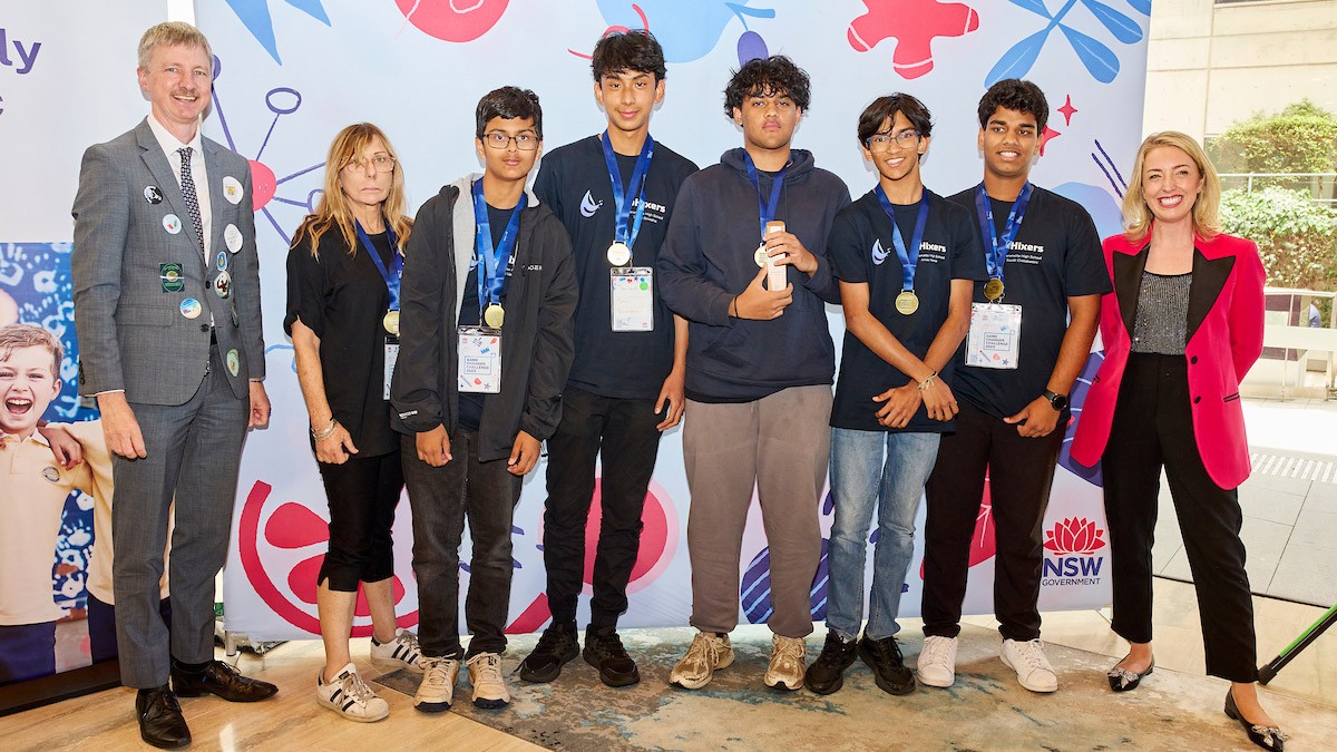 Adult male wearing grey suit stands next to adult female and five male students. A woman wearing a pink blazer stands on the side.