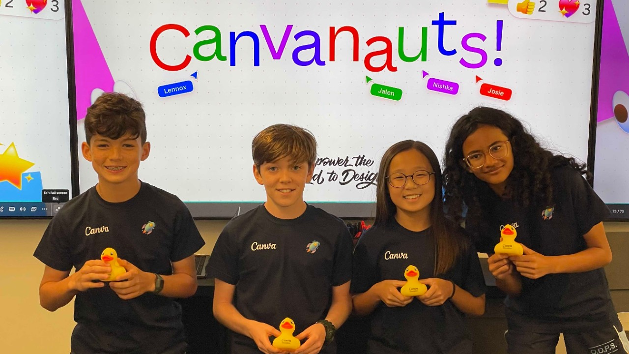 Four students stand holding yellow rubber ducks. A screen behind has the word Canvanauts written in multi-coloured text.