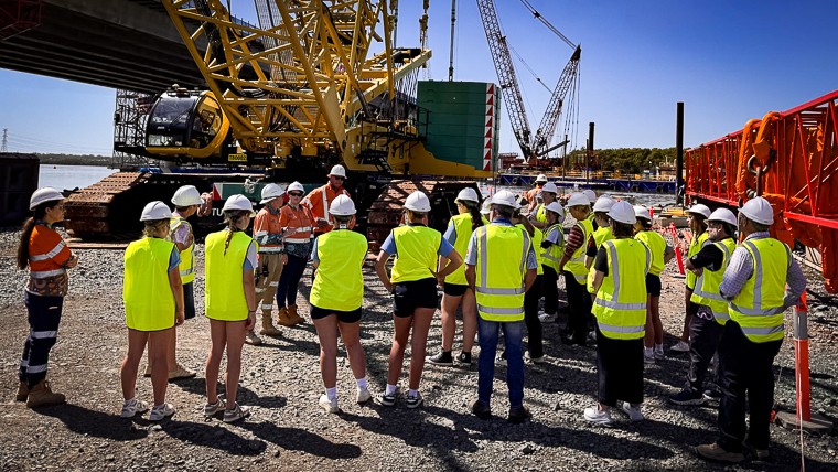 20 female studentsmin high visibility vests receiving a verbal briefing standing close by a large yellow crane.