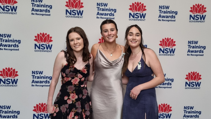 Three girls in formal dresses smiling on the red carpet entrance to the awards ceremony