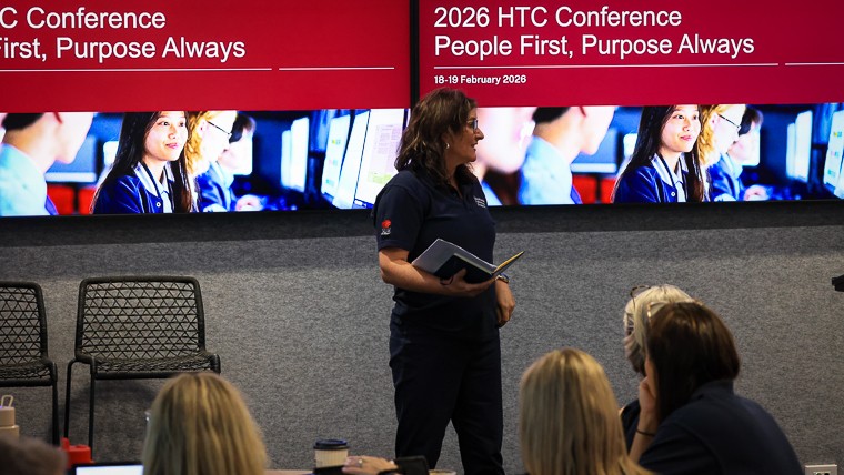Sandy Edgar EPP Career Learning Coordinator opens Day 1 of the HTC 2026 Conference with a folder in hand with 2 large screens behind with the People First Purpose Always conference theme highlighted