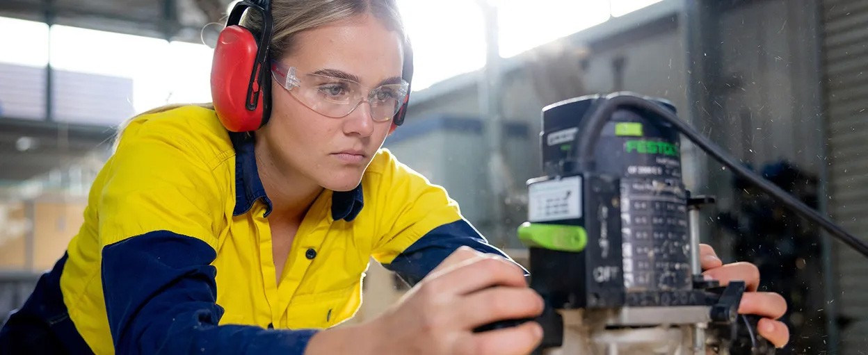 Female apprentice carpenter in safety gear with sander. 