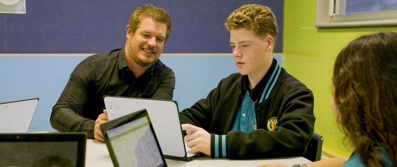 Male teacher sitting with students working on laptops