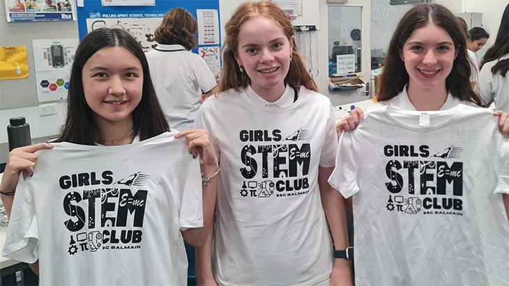Three female students holding white STEM club t-shirts with designs in black