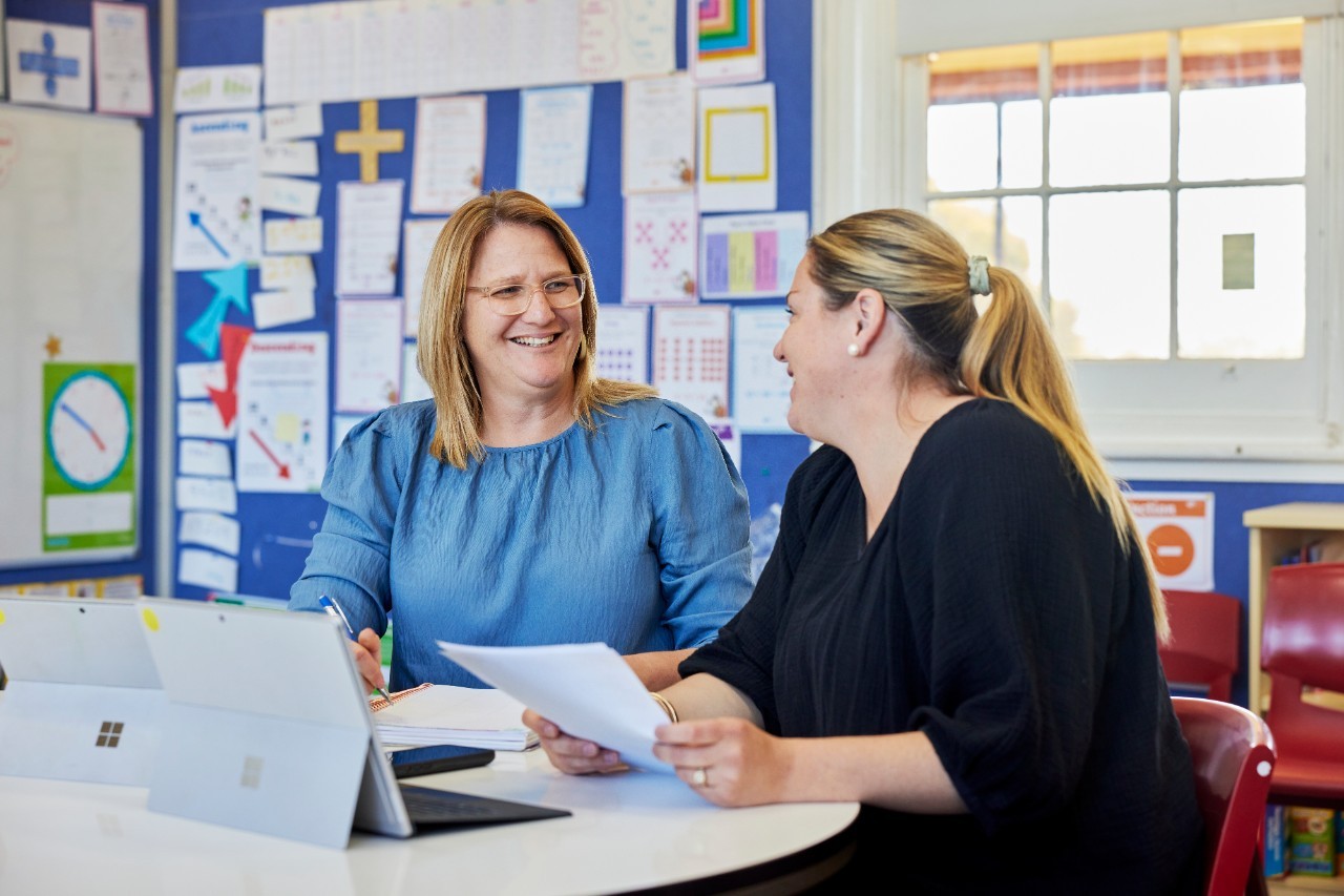 Two female educators sitting at desk. One is holding a piece of paper.