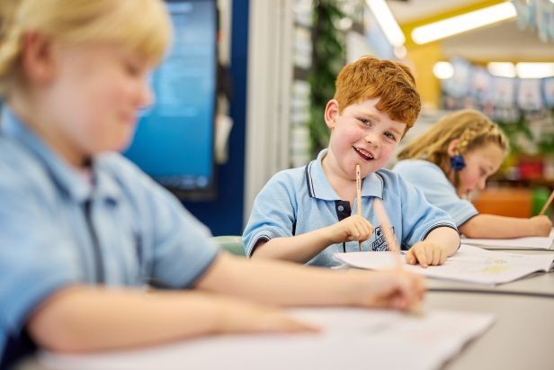 A primary school student sitting at a deak with pencil and paper smiling at the camera