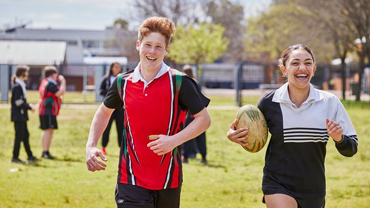 Two high school students running with a rugby ball on their school oval.
