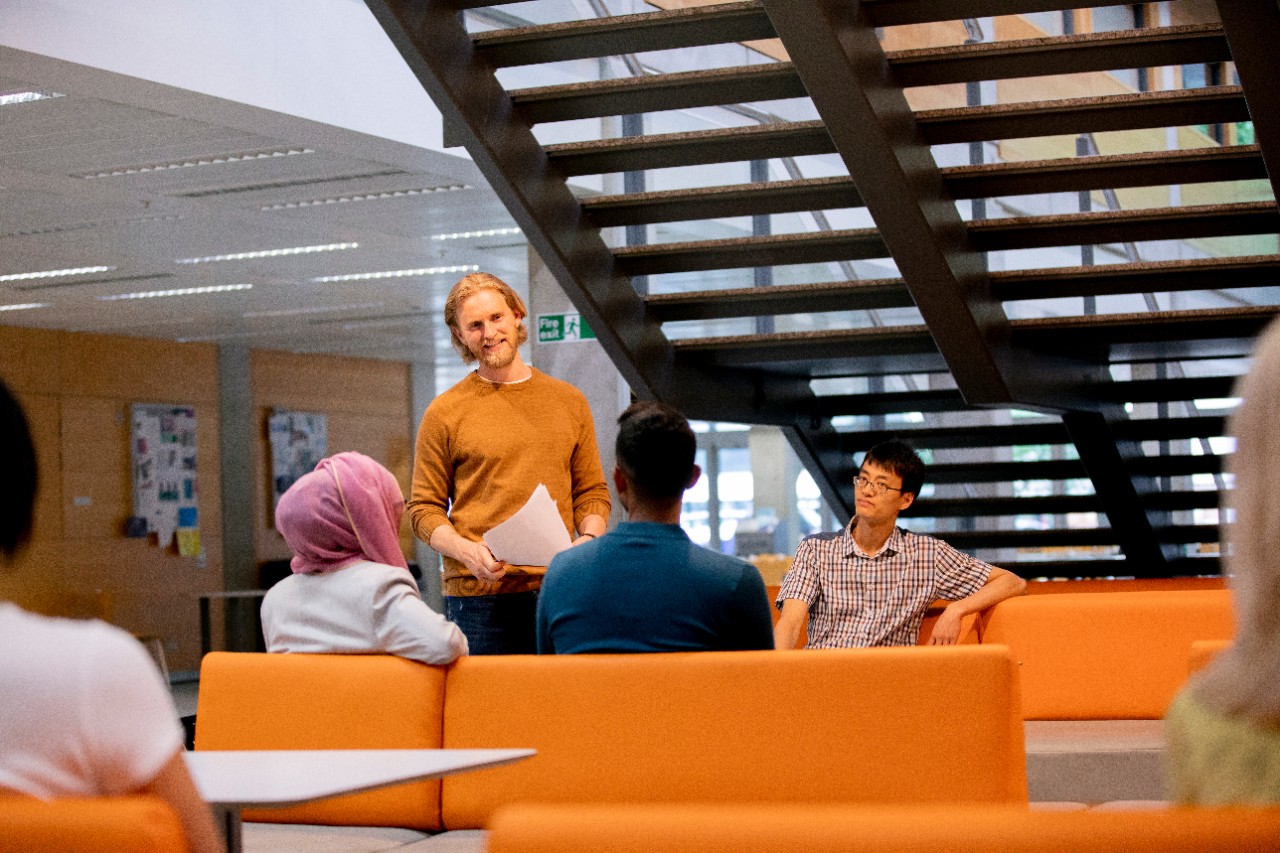 male adult holding paper speaking to one female and two male adults sitting on couch