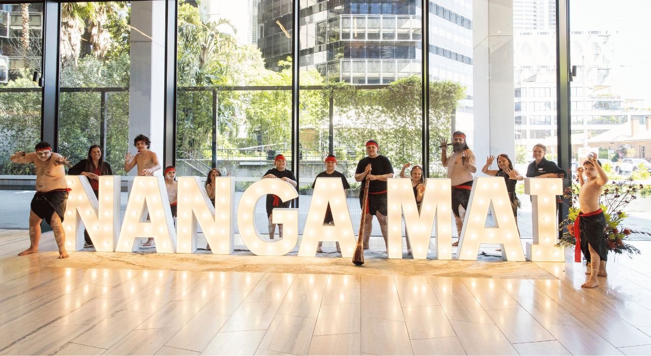 Department of Education staff and Aboriginal and or Torres Strait Islander performers posing around illuminated 3D letters spelling Nanga Mai, the events name, in a foyer with large windows and a cityscape of Parramatta in the background.