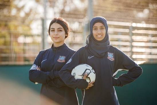 Two girls standing outside in athletic clothing, one is holding a soccer ball.