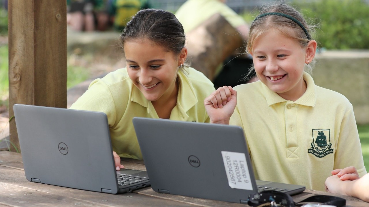 Two students sitting at an outdoor table smiling while using laptops.