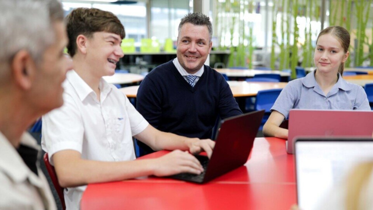 Teacher and students sitting at a desk in front of a MacBook smiling
