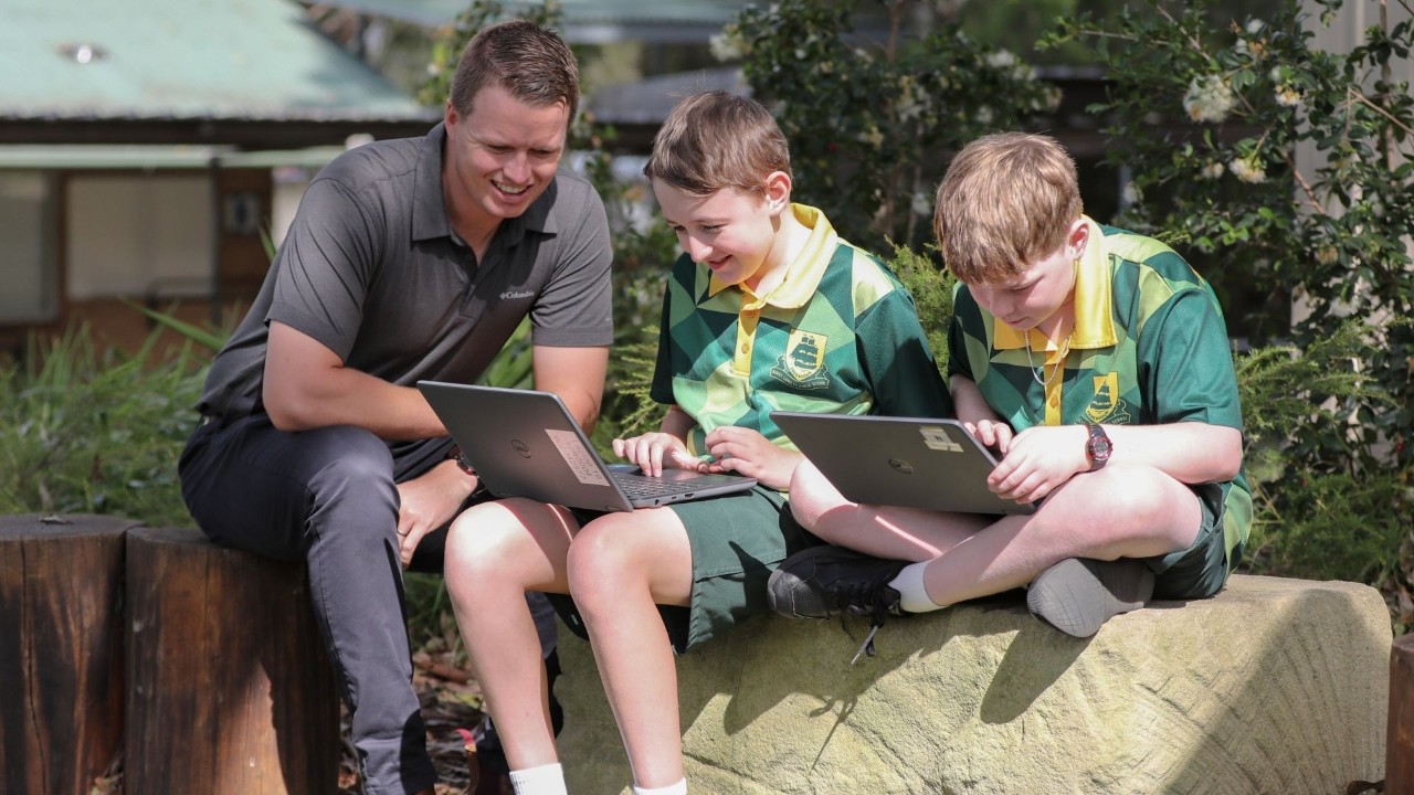 Teacher and two students sitting outside looking at a laptop.
