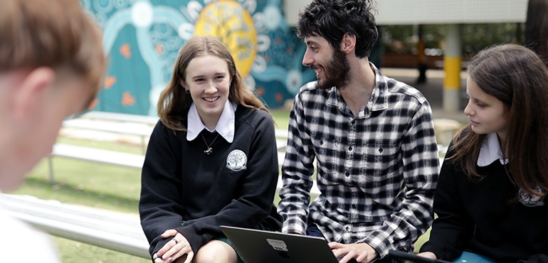 Teacher sitting outside with students with a macbook on his lap.