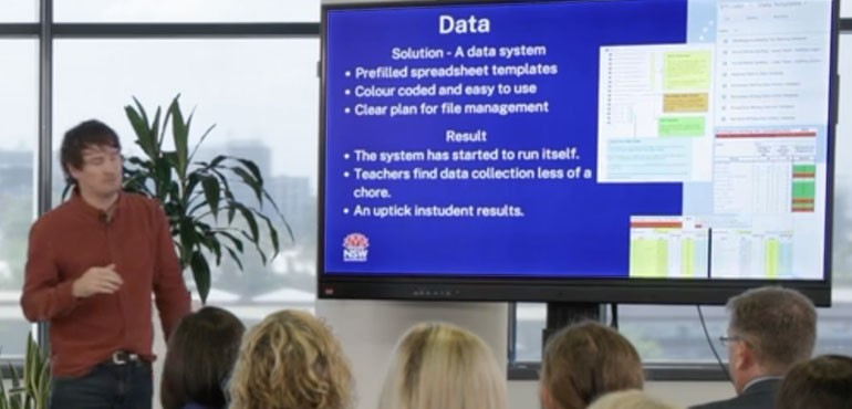 Tom giving a presentation on streamlining data collection through spreadsheet templates on a smart board.